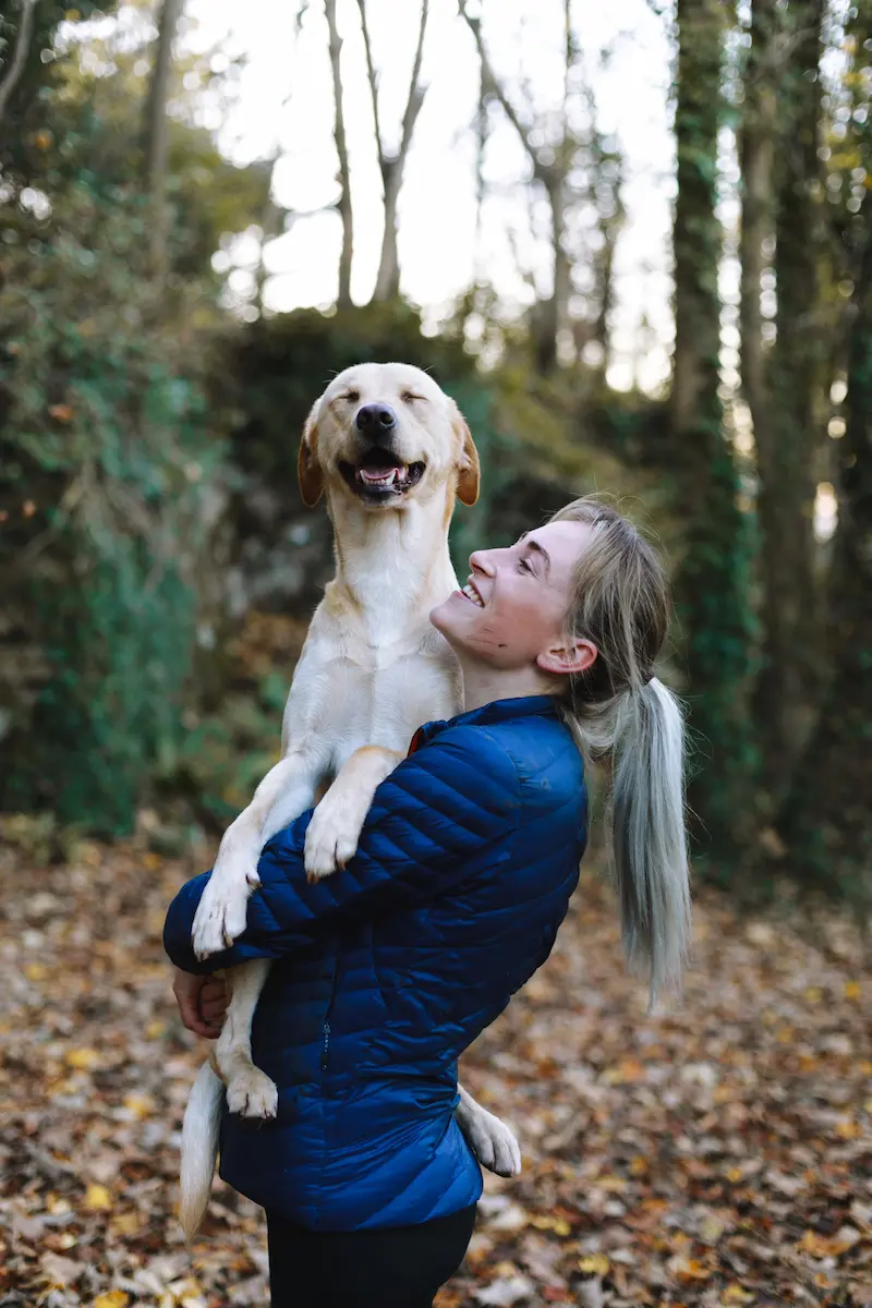 Eine Frau trägt einen lächelden, beigen Labrador in ihren Armen im Wald.